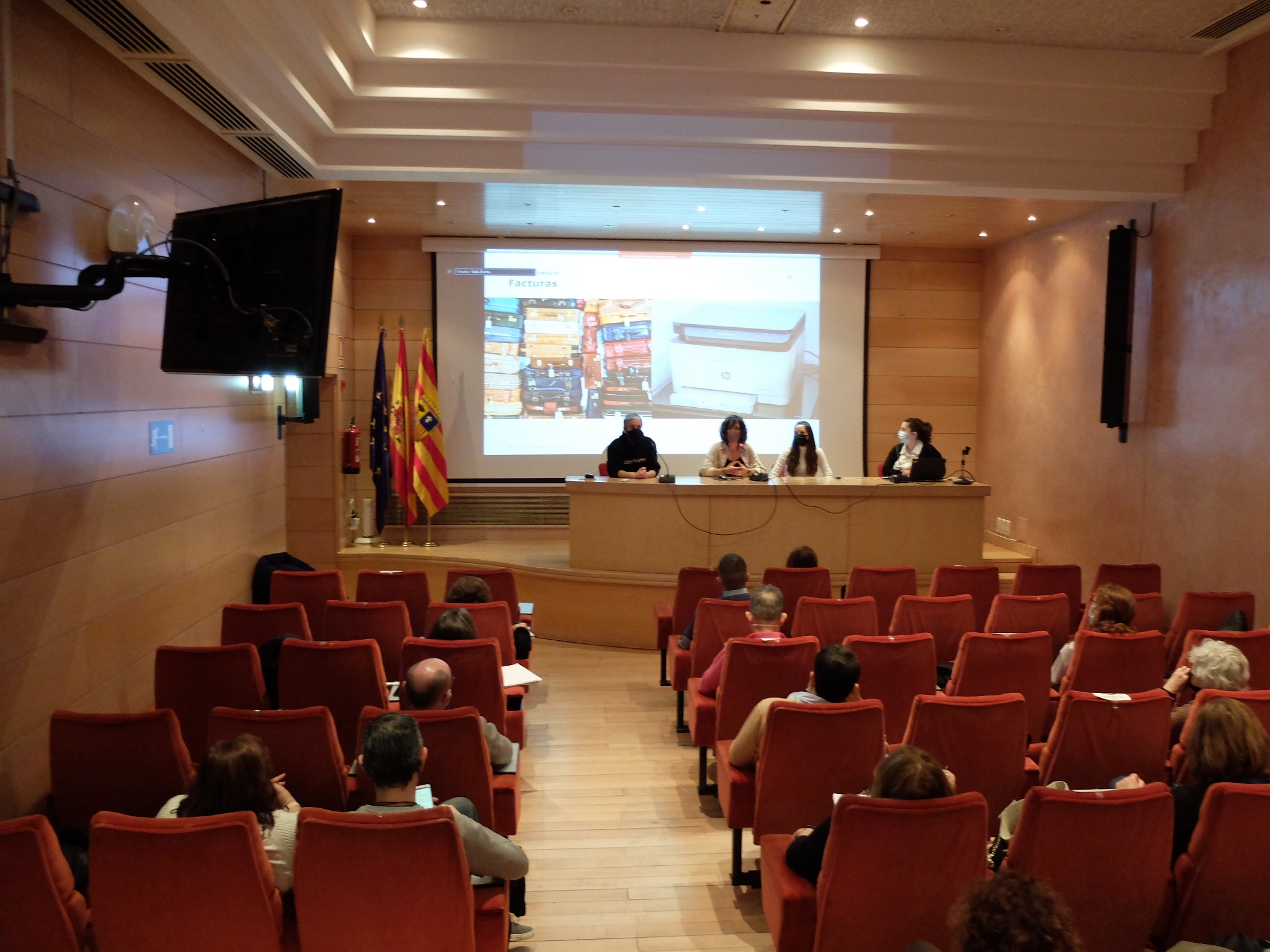 Cuatro personas en un escenario haciendo una presentación a una docena de personas sentadas en el patio de butacas. en la sala hay banderas. 