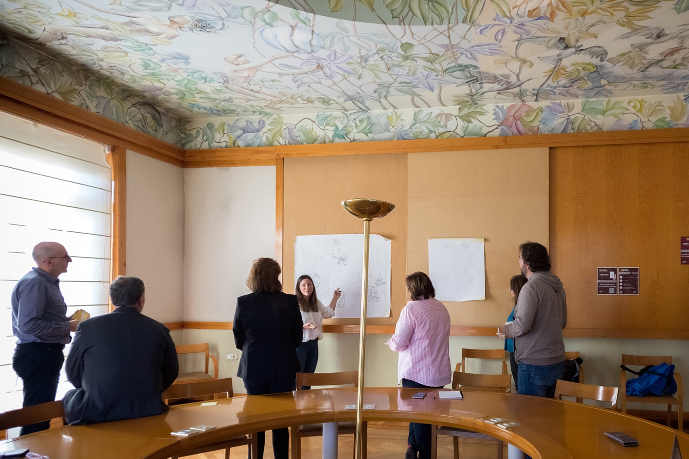 Varias personas en una sala de reuniones, escuchan a una persona en el centro que señala a un panel